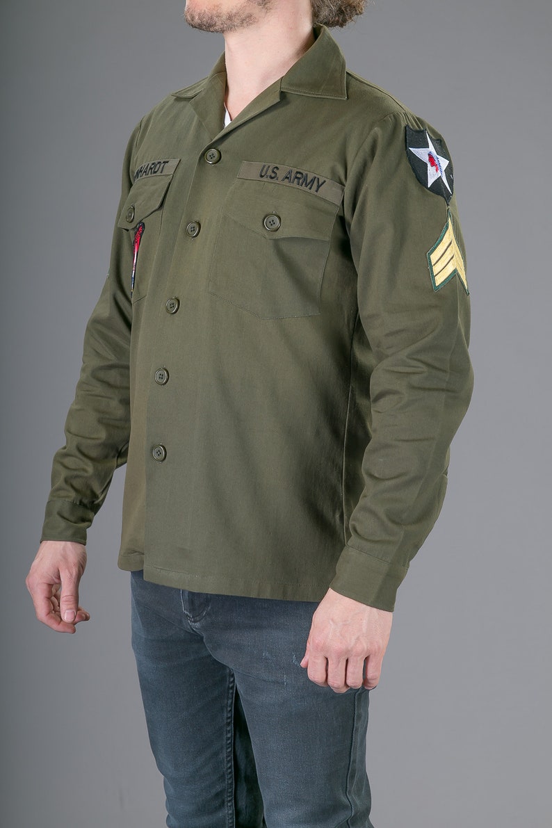 May include: A green long-sleeved shirt with a button-up collar and two pockets. The shirt has the text "US ARMY" on the left pocket and a star patch on the right sleeve.