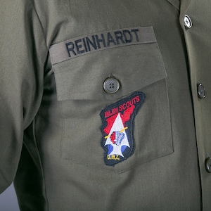 May include: A green military uniform with a name tag that reads "REINHARDT" and a patch that reads "1st.  Air  Scouts" with a red, white, and blue design.  The uniform also has a button with four holes and a patch that reads "U.S. ARMY".