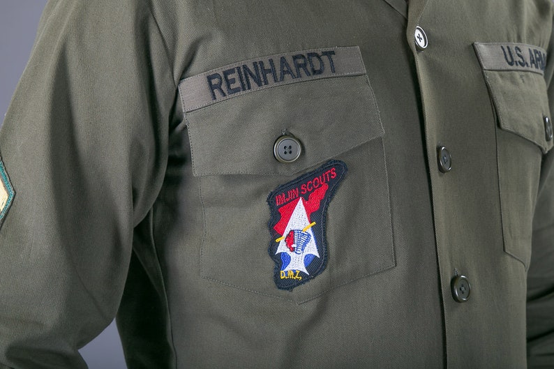 May include: A green military uniform with a name tag that reads "REINHARDT" and a patch that reads "1st.  Air  Scouts" with a red, white, and blue design.  The uniform also has a button with four holes and a patch that reads "U.S. ARMY".