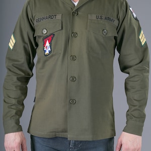 May include: A green long-sleeved shirt with a button-up front and two patch pockets. The shirt has the text "REINHARDT" and "U.S. ARMY" embroidered on the pockets. The shirt also has a patch with the text "AIR SCOUTS" and a red, white, and blue design.