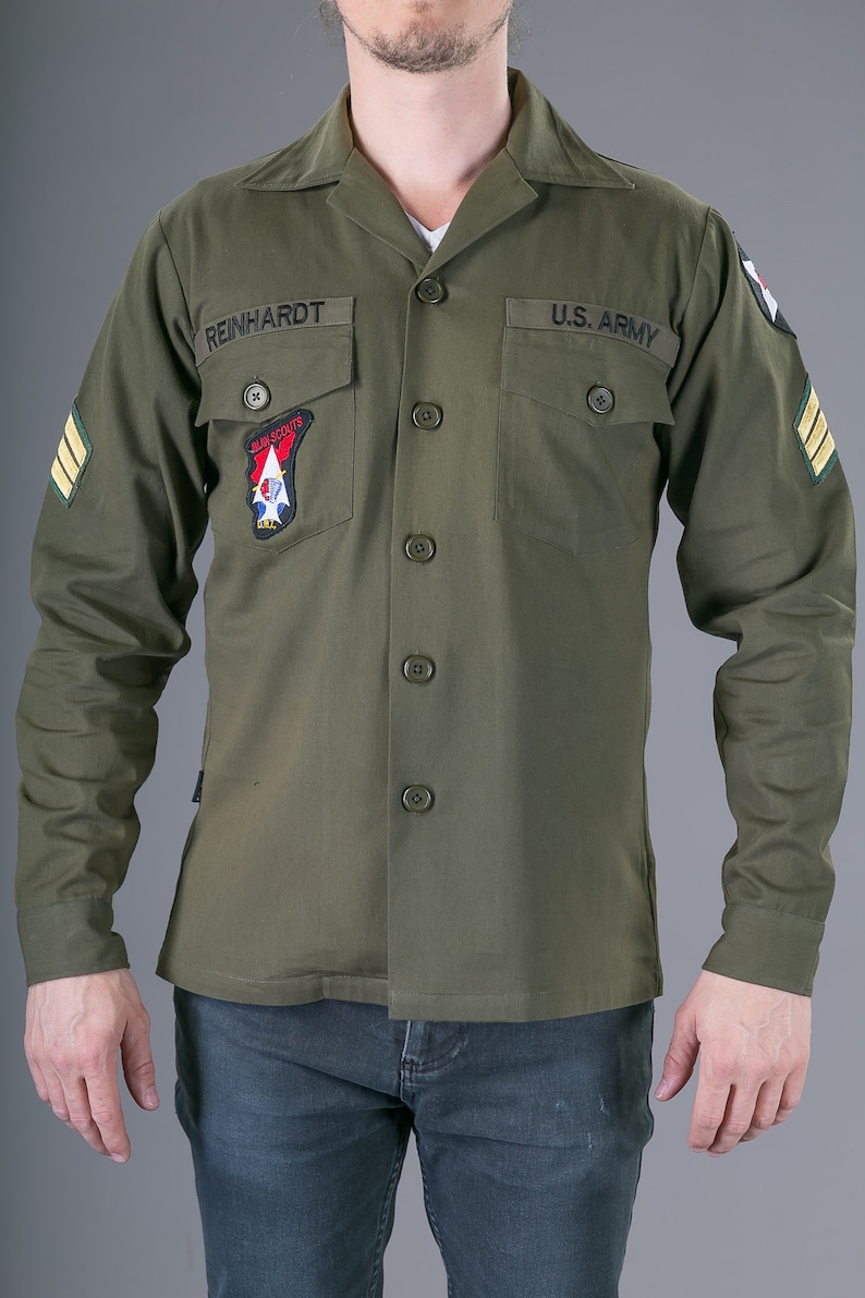 May include: A green long-sleeved shirt with a button-up front and two patch pockets. The shirt has the text "REINHARDT" and "U.S. ARMY" embroidered on the pockets. The shirt also has a patch with the text "AIR SCOUTS" and a red, white, and blue design.