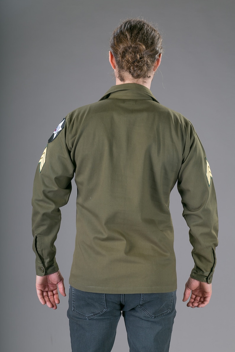 May include: A green long-sleeved shirt with a patch on the left shoulder and another on the right sleeve. The patches are yellow and green with a star and a wing design.