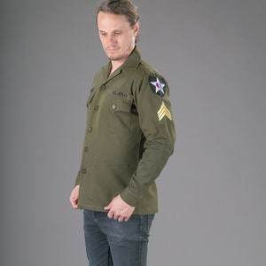 May include: A green long-sleeved shirt with a button-up front and two pockets. The shirt has a black "U.S. ARMY" patch on the left pocket and a star patch on the right shoulder.