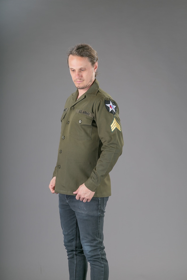 May include: A green long-sleeved shirt with a button-up front and two pockets. The shirt has a black "U.S. ARMY" patch on the left pocket and a star patch on the right shoulder.