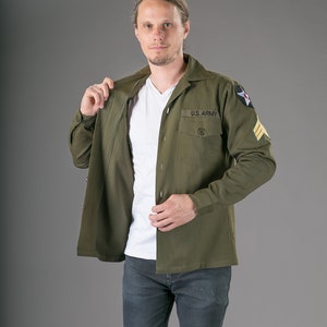 May include: A man wearing a green long-sleeved shirt with a white undershirt and blue jeans. The shirt has a patch that says "U.S. ARMY" on the left chest.