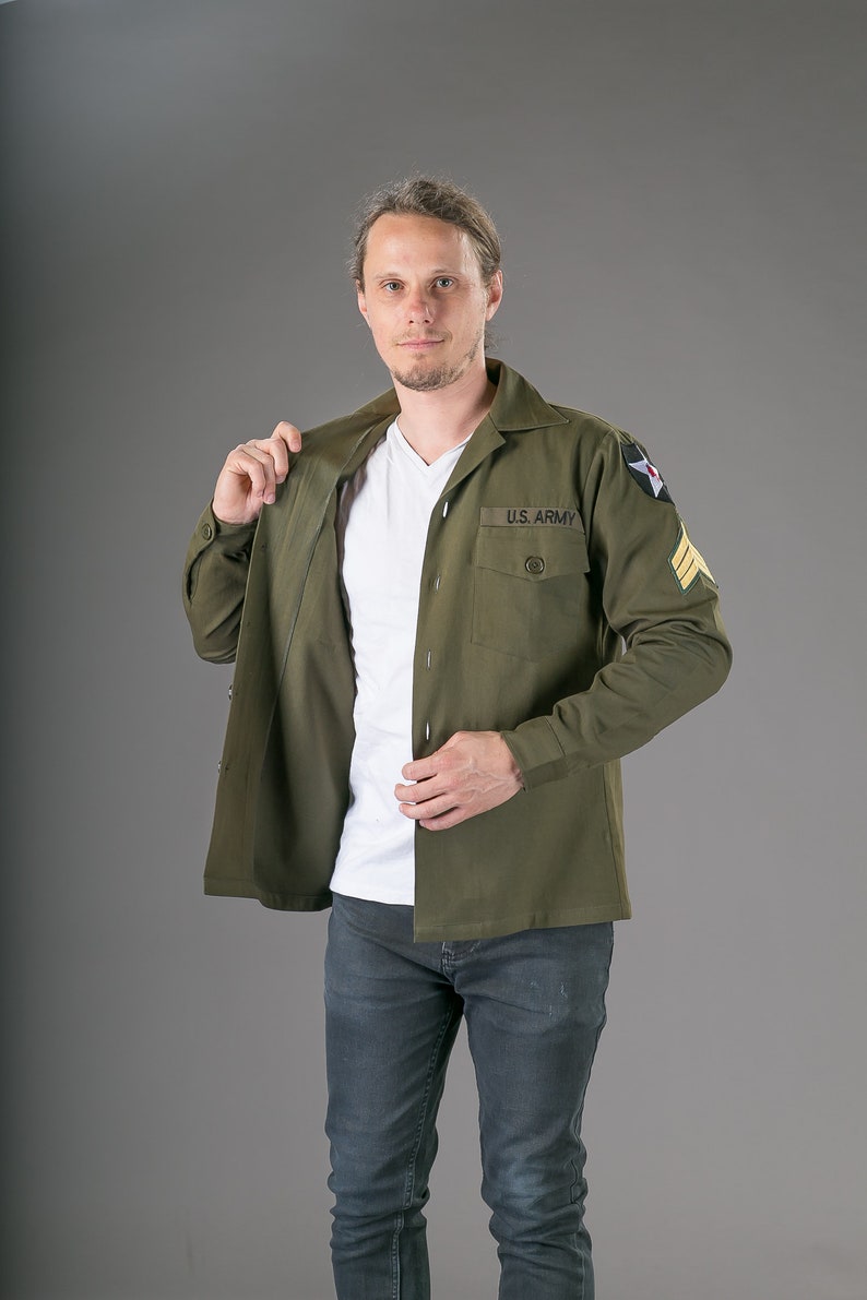 May include: A man wearing a green long-sleeved shirt with a white undershirt and blue jeans. The shirt has a patch that says "U.S. ARMY" on the left chest.