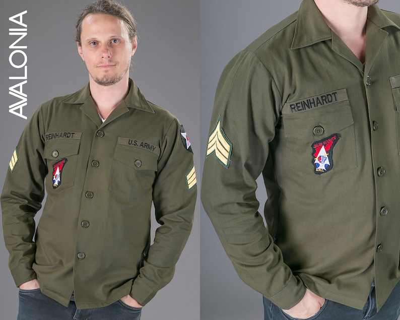 May include: A green long-sleeved shirt with a button-up collar and two pockets. The shirt has a name tag that reads "REINHARDT" and a patch that reads "U.S. ARMY".