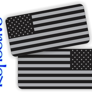 May include: Two rectangular stickers featuring a grayscale American flag design. The flag includes stars and stripes, with a reflective finish. The word "Reflective" is written vertically in blue on the left side.