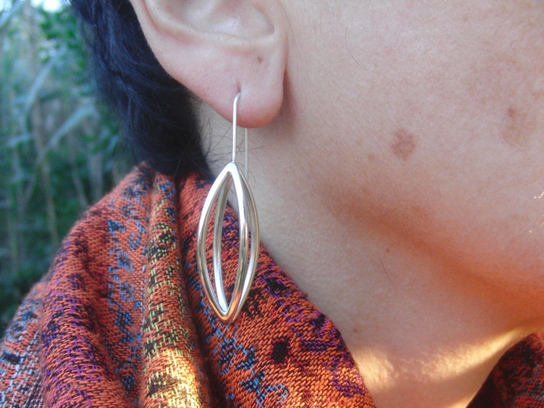 May include: Silver dangle earrings with a double oval design. The earrings are hanging from a person's ear.