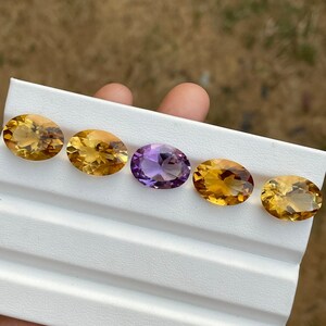 May include: Five oval-shaped gemstones displayed on a white tray. Two of the gemstones are a light golden yellow color, one is a darker golden yellow, and two are a light purple.