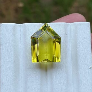 May include: A faceted, yellow-green gemstone with a pointed top and a rectangular base. The gemstone is set on a white background.