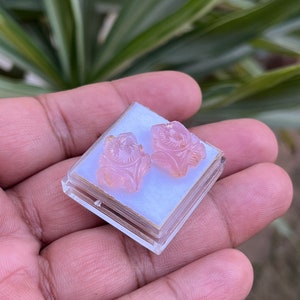 K&ouml;nnte beinhalten: Ein Paar rosa geschnitzte Rosenohrringe in einer durchsichtigen Plastikbox.