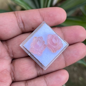 K&ouml;nnte beinhalten: Ein Paar rosa geschnitzte Blumenohrringe in einer durchsichtigen Plastikbox.