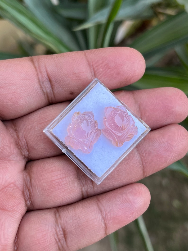 K&ouml;nnte beinhalten: Ein Paar rosa geschnitzte Blumenohrringe in einer durchsichtigen Plastikbox.