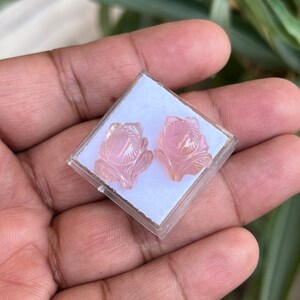 K&ouml;nnte beinhalten: Ein Paar rosafarbene, rosenf&ouml;rmige Ohrringe in einer kleinen wei&szlig;en Schachtel. Die Ohrringe sind aus einem durchscheinenden rosa Stein geschnitzt.