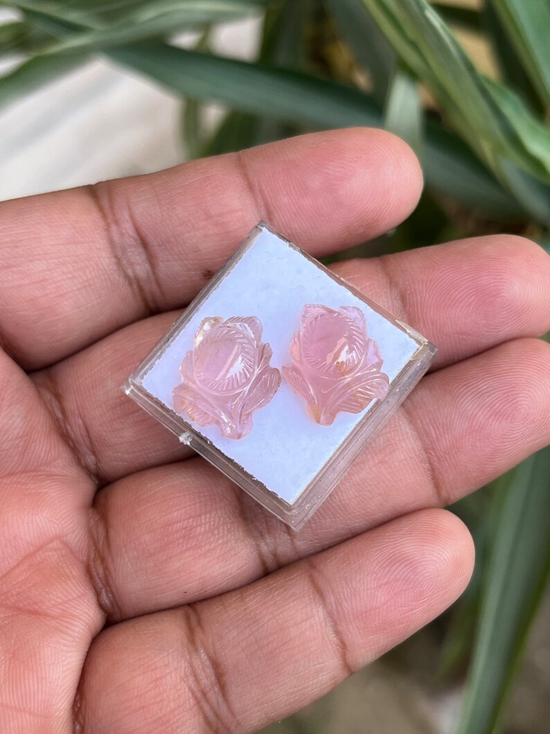 K&ouml;nnte beinhalten: Ein Paar rosafarbene, rosenf&ouml;rmige Ohrringe in einer kleinen wei&szlig;en Schachtel. Die Ohrringe sind aus einem durchscheinenden rosa Stein geschnitzt.