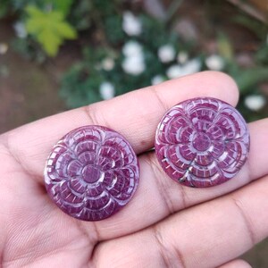 May include: Two carved ruby cabochons, each featuring a flower design. The cabochons are a deep red colour and have a polished finish.