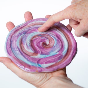 May include: A hand holding a round, pink, blue, and white ceramic coaster with a swirling pattern.