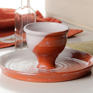 Puede incluir: Un cuenco de cerámica blanco y terracota con un esmalte blanco en el interior y un esmalte de terracota en el exterior. El cuenco está sentado sobre un plato de terracota con un esmalte blanco en el interior.