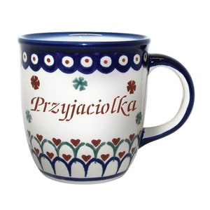 May include: A white ceramic mug with a blue rim and handle. The mug has a floral pattern in red, green, and blue. The words "Przyjaciółka" are written in brown on the front of the mug.