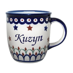 May include: A white ceramic mug with a blue rim and handle. The mug has a blue and white pattern around the top, red stars and a blue star pattern in the middle, and a green and red floral pattern around the bottom. The word "Kuzyn" is written in black cursive on the front of the mug.