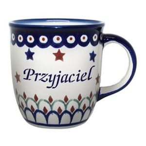 May include: A white ceramic mug with a blue rim and handle. The mug has a blue and red star pattern and the word "Przyjaciel" written in black.