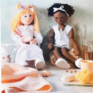 May include: Two handmade cloth dolls, one blonde and one with dark hair, are seated. The blonde doll wears a white dress and the other doll wears a white top and shorts. The dolls are on a shelf with bath accessories.