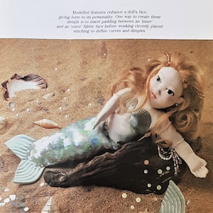 May include: A mermaid doll with long, curly red hair, a white face, and blue eyes. She is wearing a green and blue sequined tail and is lying on a piece of driftwood. The doll is surrounded by seashells and sand.