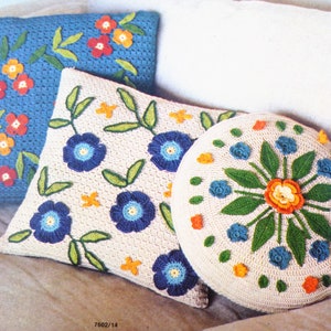 May include: Three crocheted cushions with floral designs. The cushions are white with blue, green, orange, and yellow flowers. The cushions are on a white background.