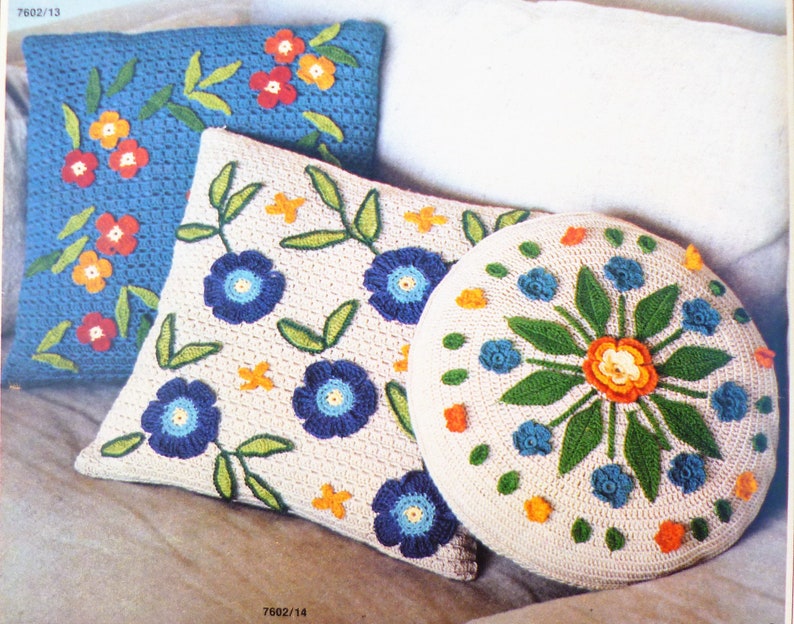 May include: Three crocheted cushions with floral designs. The cushions are white with blue, green, orange, and yellow flowers. The cushions are on a white background.