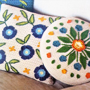 May include: Two crocheted cushions with floral designs. One cushion is square and the other is round. Both cushions have a cream background with blue, green, orange, and yellow flowers.