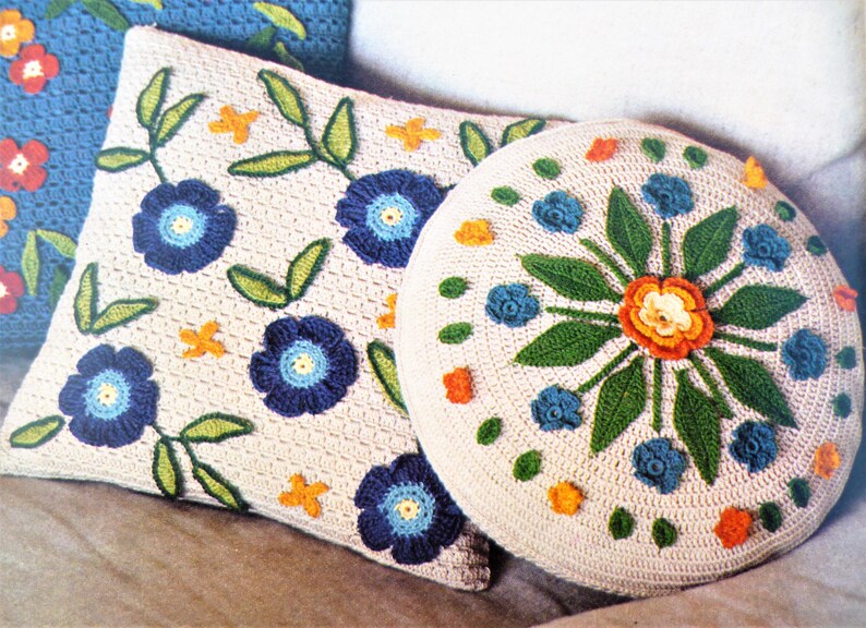 May include: Two crocheted cushions with floral designs. One cushion is square and the other is round. Both cushions have a cream background with blue, green, orange, and yellow flowers.