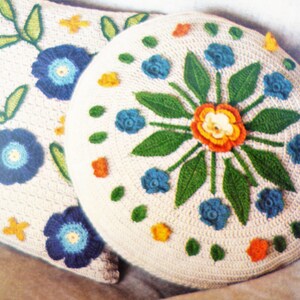May include: Two crocheted cushions featuring floral designs. The larger cushion has a central flower with orange, yellow, and white petals surrounded by green leaves and blue flowers. The smaller cushion has a similar design with blue flowers and green leaves.