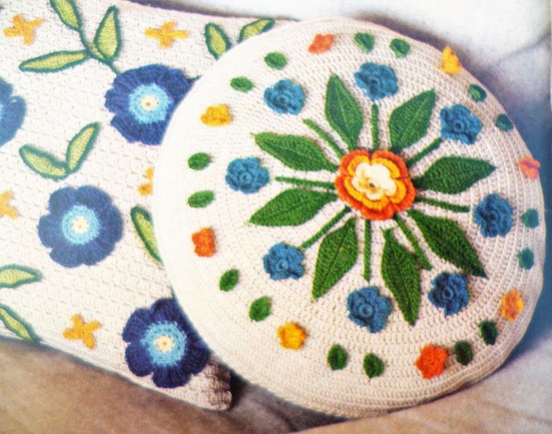 May include: Two crocheted cushions featuring floral designs. The larger cushion has a central flower with orange, yellow, and white petals surrounded by green leaves and blue flowers. The smaller cushion has a similar design with blue flowers and green leaves.