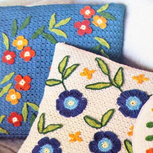 May include: Three crocheted cushions featuring floral designs. The cushions are made with white, blue and green yarn. The flowers are red, orange and blue.