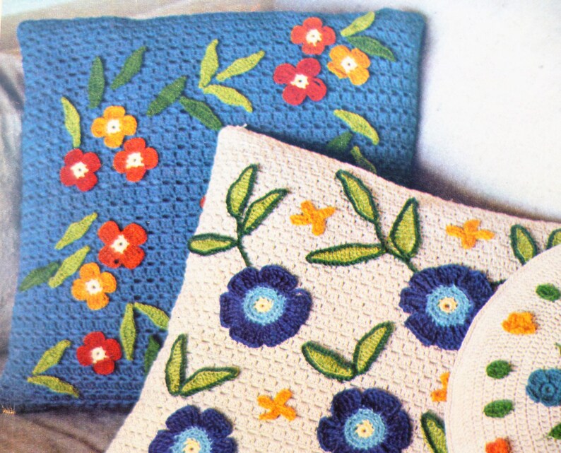 May include: Three crocheted cushions featuring floral designs. The cushions are made with white, blue and green yarn. The flowers are red, orange and blue.