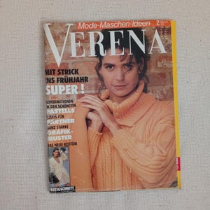 Op de afbeelding: Een vintage editie van het tijdschrift Verena uit februari 1992 met een persoon in een perzikkleurige kabeltrui met een hoge kraag en knopen. De cover bevat Duitse tekst zoals Mode-Maschen-Ideen en Mit Strick ins Frühjahr.