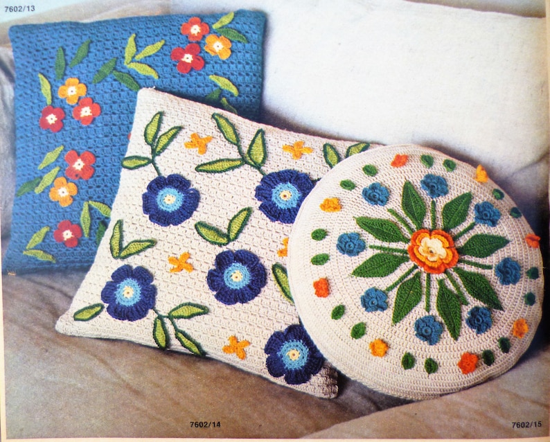 May include: Three crocheted cushions with floral designs. The cushions are white with blue, orange, and green flowers. The cushions are on a white background.