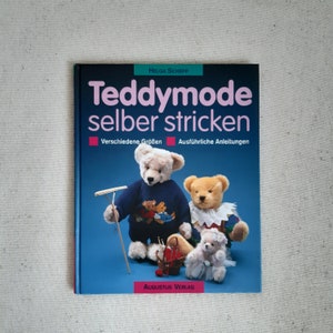 Puede incluir: Un libro titulado "Teddymode selber stricken" de Helga Schepp. La portada del libro presenta tres osos de peluche, uno con un suéter azul, otro con un suéter blanco y un oso de peluche más pequeño con un vestido blanco. La portada del libro es azul con acentos rojos y rosados. El libro está publicado por Augustus Verlag.