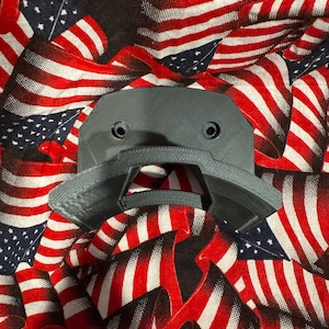 May include: A gray, 3D-printed object with two circular holes and a curved design, set against a backdrop of American flags in red, white, and blue. The object has a textured surface and a geometric shape.