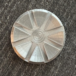 May include: A silver-colored, circular object with a textured surface. The design features a central circle surrounded by radiating, wedge-shaped segments, each with concentric lines. The object appears to be made of metal.