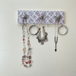 May include: A white and grey patterned wooden board with five hooks and three necklaces hanging from them. The necklaces are made of beads, metal, and chain. The board has a decorative design with a geometric pattern.