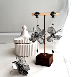 May include: A set of grey flower earrings and a matching ring are displayed on a gold jewellery stand. The earrings have dangling chains with black beads. A white, lidded container is in the background.