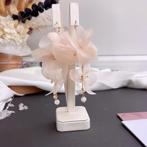 Pode incluir: Um par de delicados brincos de flores cor de rosa claro com detalhes de cristal pendurados em uma corrente dourada.