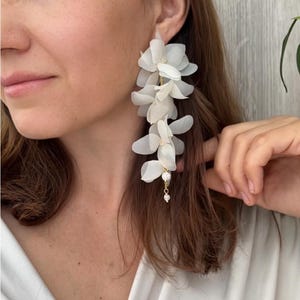 Op de afbeelding: Witte bloemen oorbellen met meerdere lagen stoffen bloemen en goudkleurige accenten. De oorbellen hebben een delicaat, trapsgewijs ontwerp, met kleine, bungelende versieringen.
