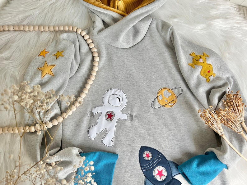May include: A gray hooded sweatshirt with embroidered astronaut, planet, and stars. The sweatshirt has a yellow hood lining and blue sleeves.