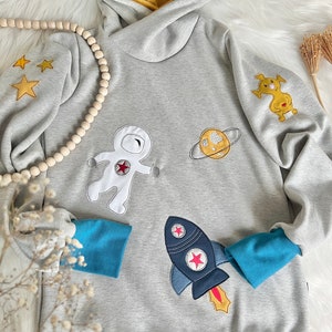 May include: A gray hooded sweatshirt with a blue and yellow trim. The sweatshirt features embroidered astronaut, rocket, planet, and star designs.