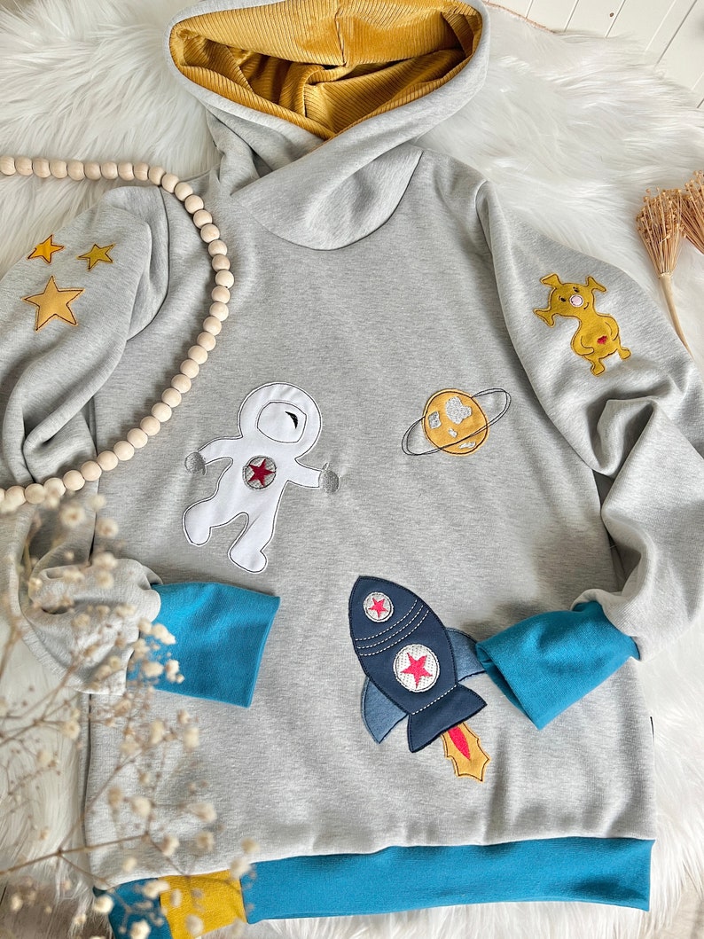 May include: A gray hooded sweatshirt with a blue and yellow trim. The sweatshirt features embroidered astronaut, rocket, planet, and star designs.