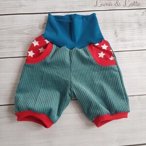 Puede incluir: Pantalones cortos de beb&eacute; de pana color turquesa con ribetes y bolsillos rojos. Los bolsillos tienen un borde rojo con detalles de estrellas blancas. La cintura es azul y los pantalones cortos se muestran sobre una superficie de madera clara. El texto "Luna & Lotte" es visible.