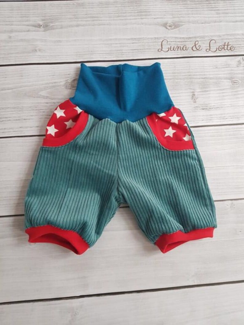 Puede incluir: Pantalones cortos de beb&eacute; de pana color turquesa con ribetes y bolsillos rojos. Los bolsillos tienen un borde rojo con detalles de estrellas blancas. La cintura es azul y los pantalones cortos se muestran sobre una superficie de madera clara. El texto "Luna & Lotte" es visible.